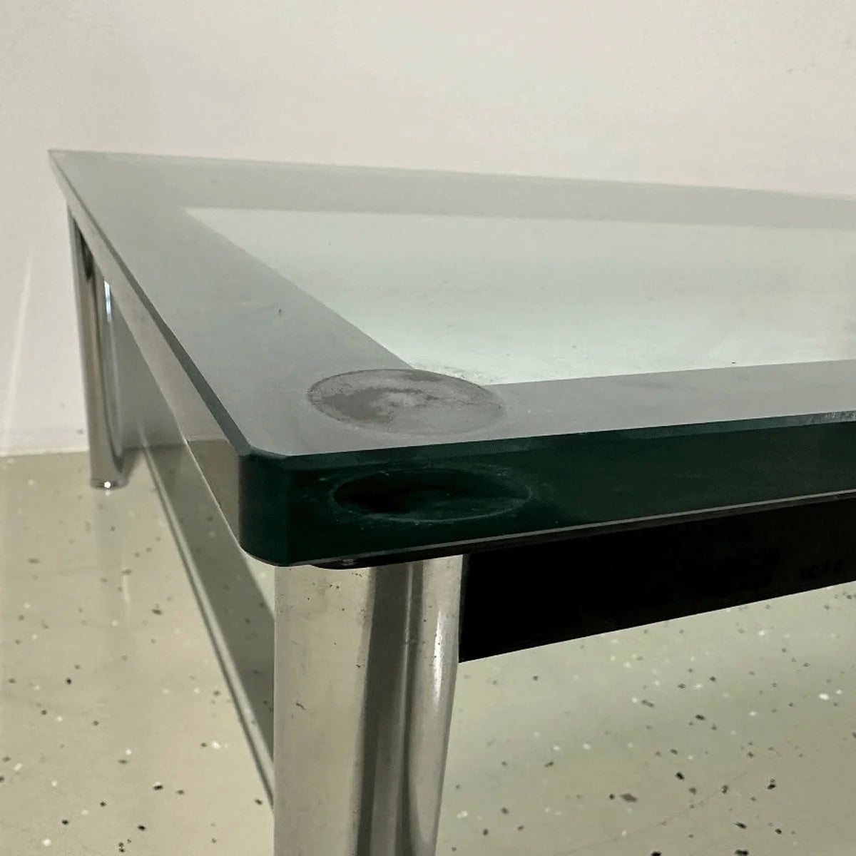 Kante der Glasplatte und Übergang zum schwarzen Rahmen am Cassina LC10-P, verchromtes Bein im Hintergrund.