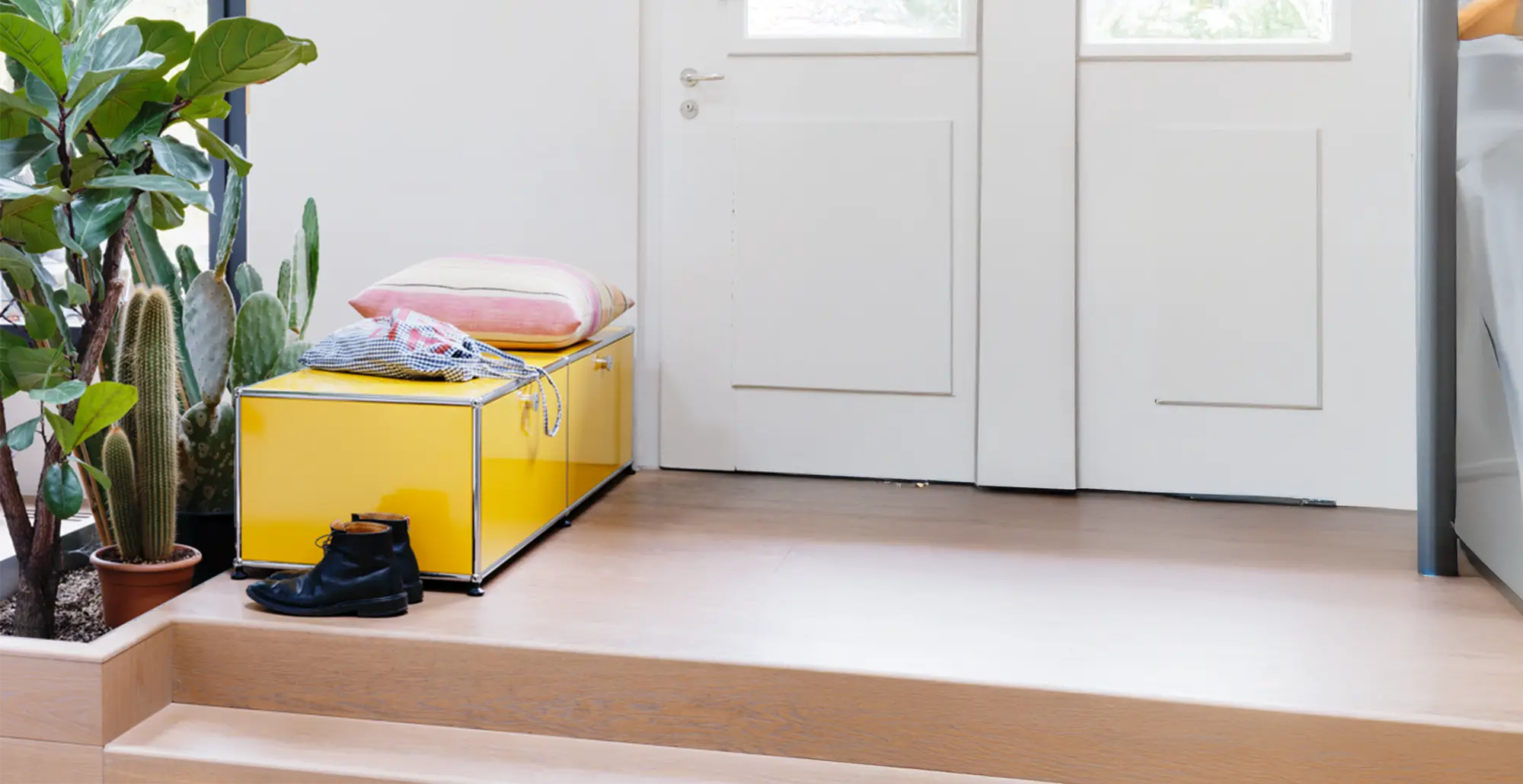 Gelbes USM Haller Lowboard als Sitzbank im Flur mit Kissen und Kleidung, daneben Pflanzen und Schuhe vor weisser Tuer
